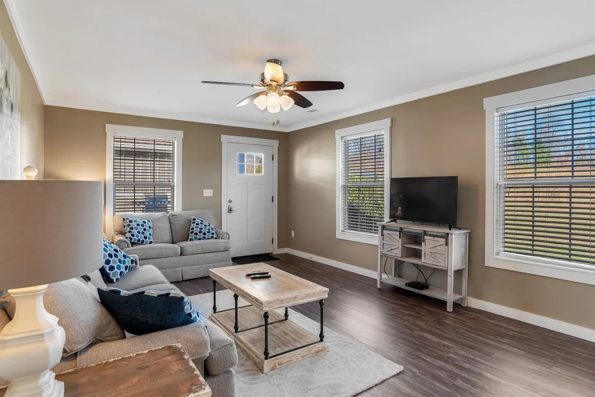 A stylish living area featuring a cozy sofa arranged around a modern coffee table, a sleek television set, and an elegant light fixture overhead. The room is awash in natural light, with soothing decor tones creating a tranquil, welcoming atmosphere.