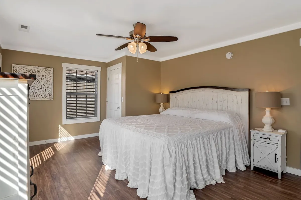 A serene bedroom with a bed dressed in crisp white linens, framed by soft decorative lamps and tasteful artwork. Gentle natural light streams in, enhancing the room's peaceful and inviting mood.