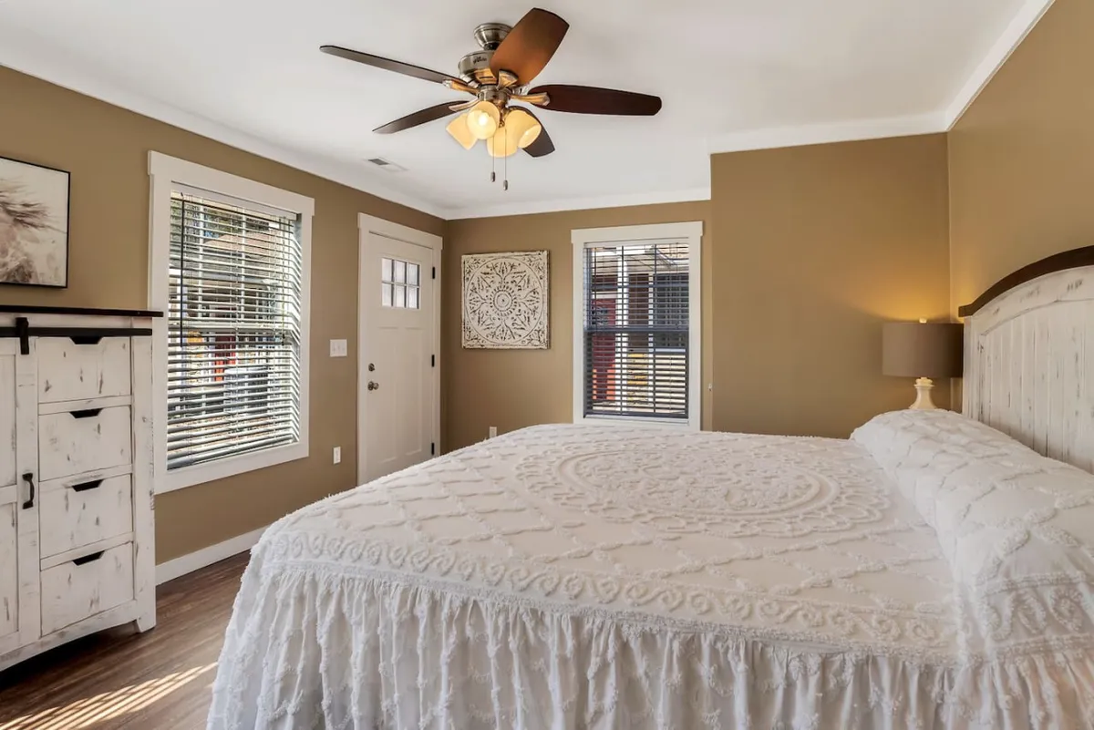 The bedroom is depicted with an intricately made king bed, a classic wooden dresser, ceiling fan, and door with an inset window. The space feels warm and welcoming, ideal for relaxing, quiet moments, or starting the morning gently.
