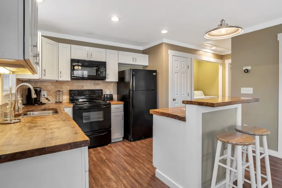 This image showcases a well-appointed kitchen with a prominent island, sleek black appliances, wooden countertops, and pristine white cabinetry. The ambiance is welcoming, with visible space for cooking, entertaining, and casual dining. The layout encourages interaction and connection with the adjoining living area.