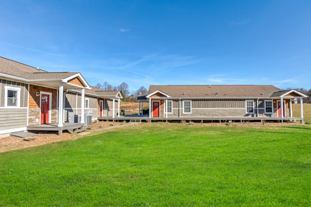 This exterior shot features welcoming red doors and wooden porches set against a lush green lawn and vibrant blue sky. The scene hints at gatherings, outdoor dining, and relaxation, showcasing a friendly, approachable atmosphere.