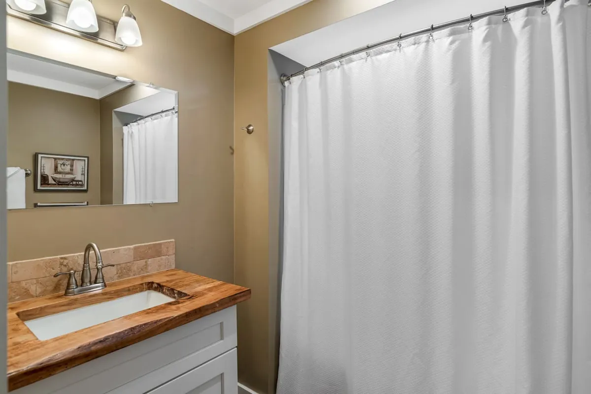 A bright, modern bathroom featuring a well-lit vanity with a spacious sink, a mirror, and a clean shower area partially visible behind a curtain. Soft lighting and neutral colors create a serene atmosphere, emphasizing the sense of cleanliness and relaxation. Fluffy towels are neatly arranged, and the space is uncluttered, reinforcing a spa-like environment.