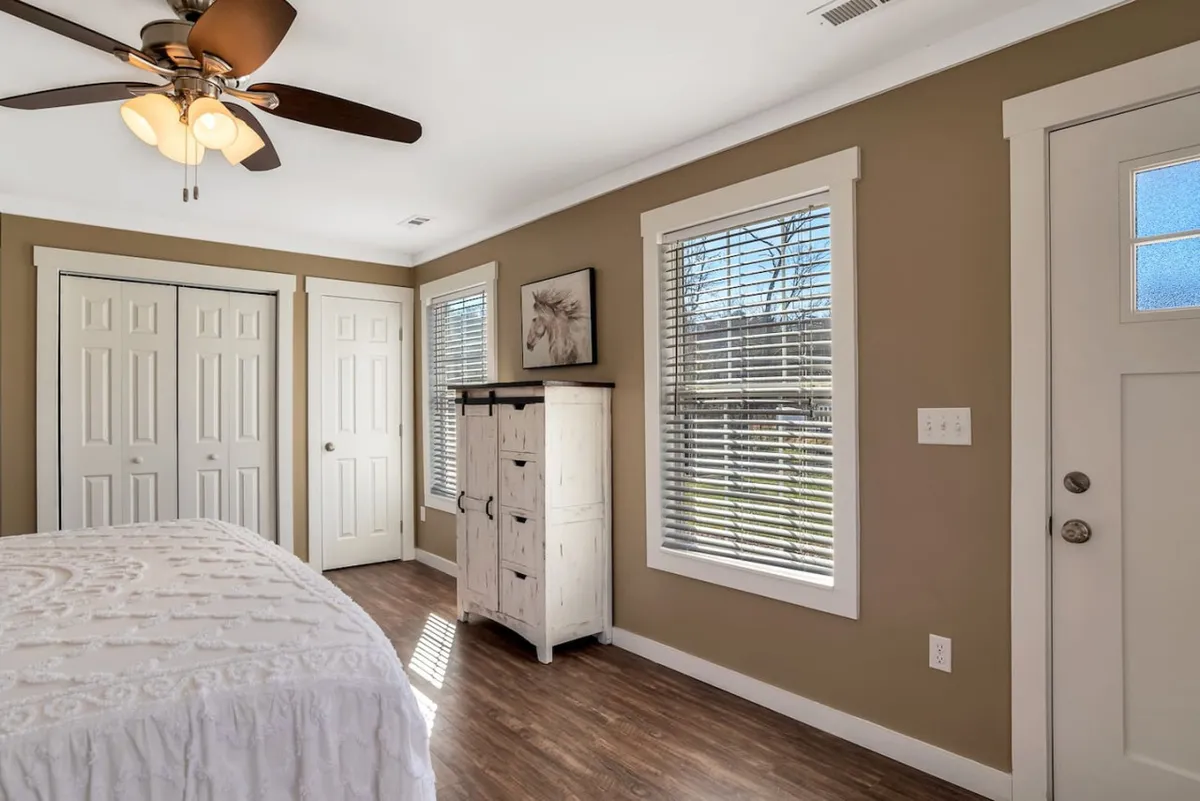 This image presents a bright and inviting bedroom with a king bed, a sturdy dresser, large windows, and a door leading to another part of the home. Soft natural light washes over the space, revealing crisp linens and a tranquil atmosphere perfect for rest or reading.